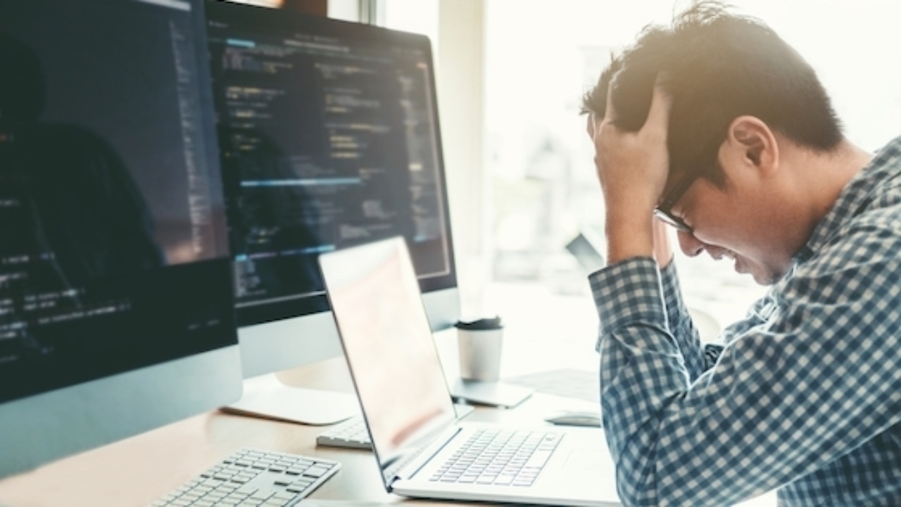 Programming is difficult, make sure to manage your stress. A programmer sitting in front of three computers holding his head in his hands in frustration.